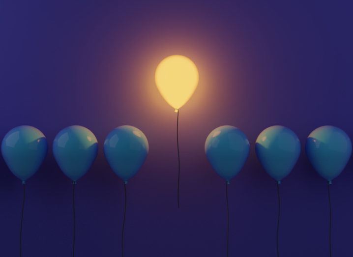 A glowing yellow balloon floats higher than a line of dark blue balloons against a purple background.