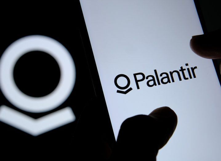 Silhouette of fingers on a smartphone, which has the Palantir logo on the screen.