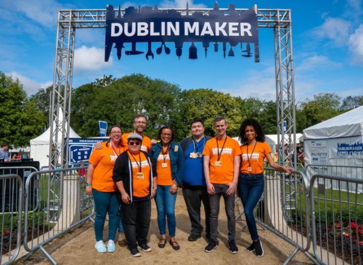 A group of people in matching orange Dublin Maker T-shirts pictured in a park on a sunny day. A sign above their head bears the Dublin Maker logo.