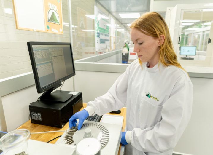 A scientist in a lab coat is working to analyse a soil and sediment sample in the new geological facility. They are placing soil samples into the analysis device, which is an off-white-coloured box and is connected to a computer monitor that is on the same desk.