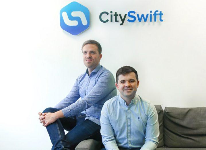 Two men are sitting on a couch beneath a sign that says 'CitySwift'.