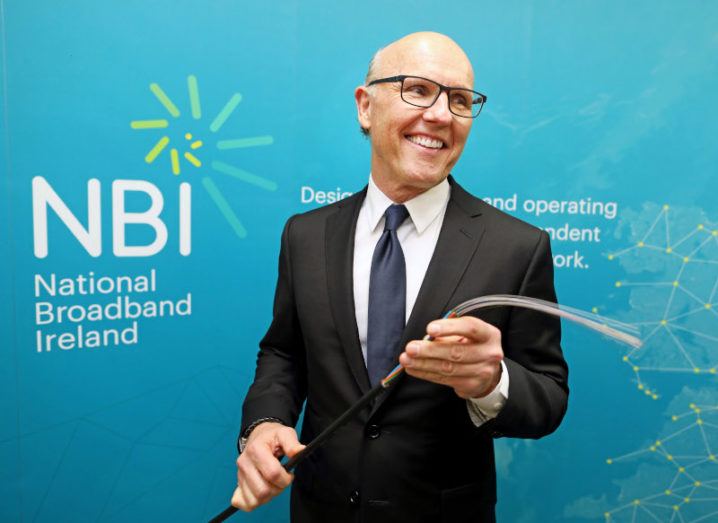 David McCourt stands in a suit, holding a fibre cable, in front of a sign that says National Broadband Ireland.