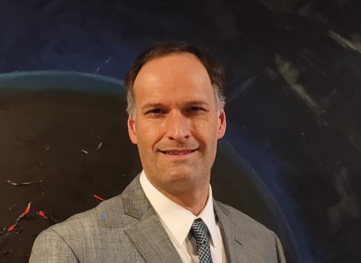 A man in a grey suit and patterned tie pictured against an abstract painting of a planet in space.