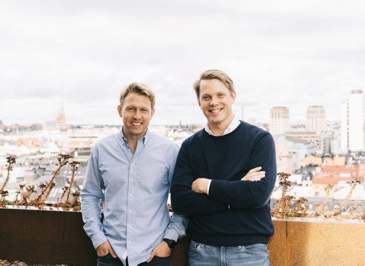 Two men stand on a rooftop with a city behind them.