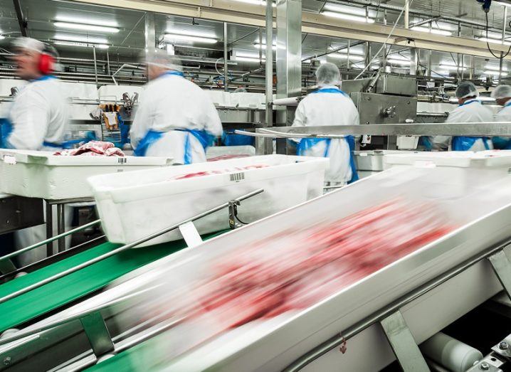 Meat whizzes down a production line in a factory, with workers in protective gear working in the background.