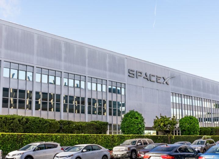 The SpaceX building is pictured from the parking lot. Cars are visible in front of the building. The SpaceX logo is on the side, with windows built into the tall, grey walls.