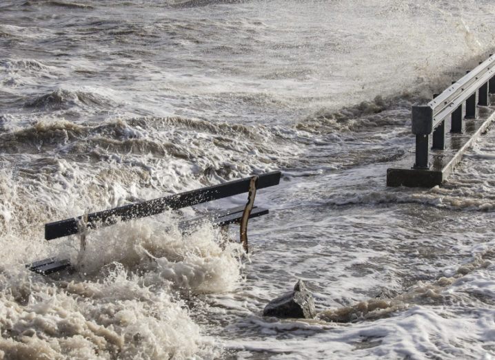 A flood surges over a bench next to a road.