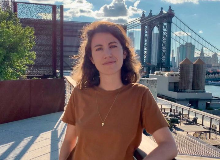 A woman leans on a railing on a sunny day against a cityscape.