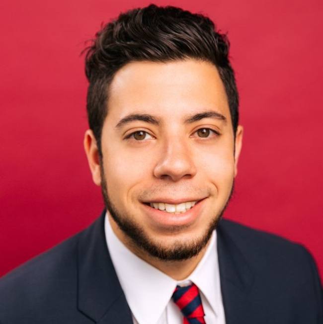 A close-up headshot of a man in a suit against a red background.