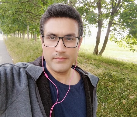 A selfie of a man out on a walk among some greenery. He wears casual clothes and has earphones in his ears.