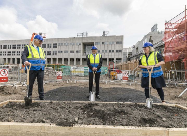 Three people in high-vis vests and hard hats stand with shovels at a building site on the Trinity College Dublin campus.