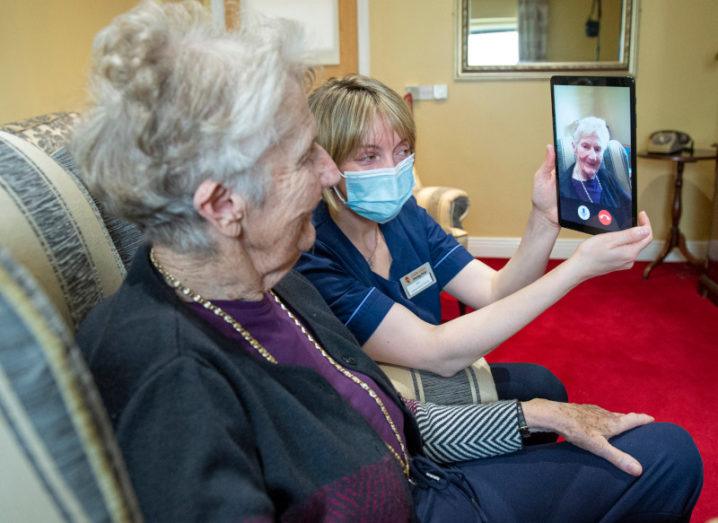 A woman in a nursing home sits in a chair while a nurse wearing a face covering holds up a tablet showing the face of another woman.