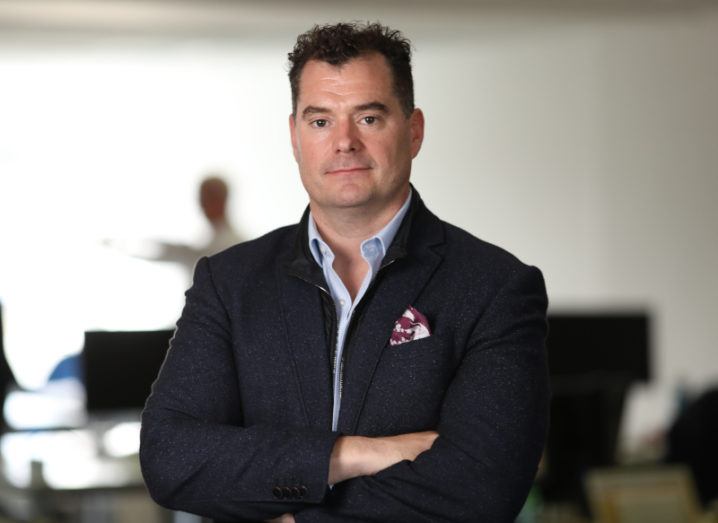 A man in a navy wool blazer with a wine-coloured pocket square and powder blue shirt stands with his arms folded in an office setting.