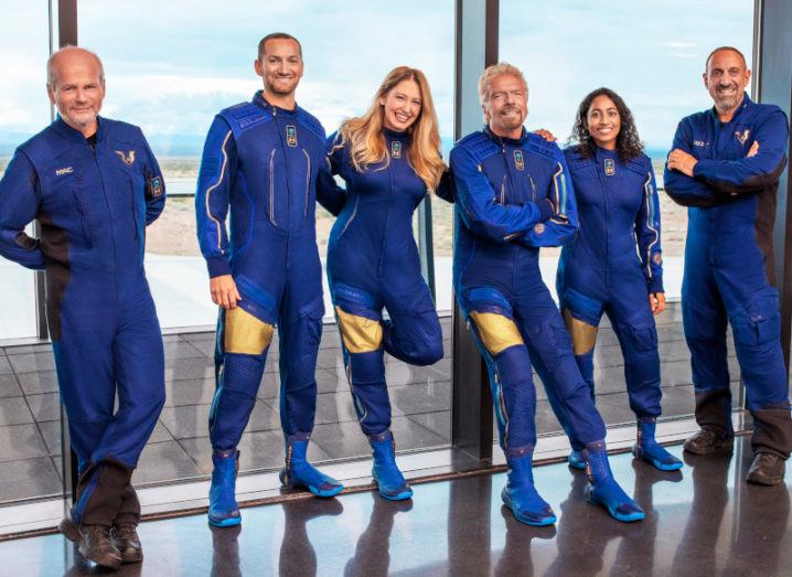 The crew of the VSS Unity are pictured standing and smiling. They are all wearing identical blue jumpsuits and are leaning against a glass wall. A runway can be seen through the glass.