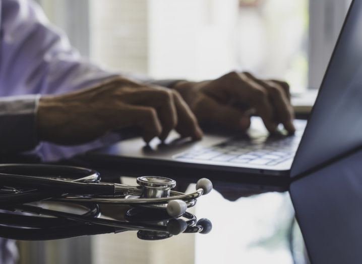 A man with a doctor’s coat sits at a table working on a laptop while a stethoscope sits beside him.