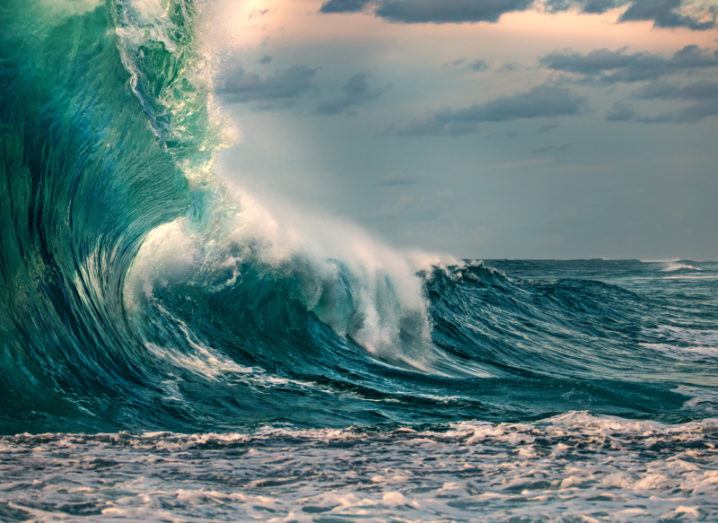 A large wave in the ocean is depicted. There is some white froth, but most of the water is a dark blue with a green hue. The sky is a light grey with hints of orange and the occasional cloud.