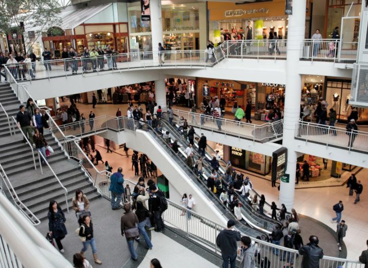 A busy shopping centre is pictured. Three stories of the shopping centre are visible and there are people outside of all the shops. There is an escalator and a staircase going between each of the levels.