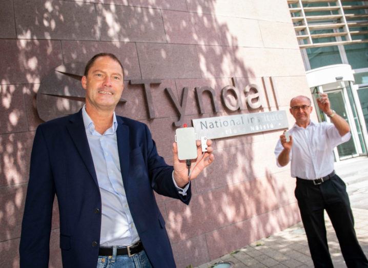 Mike Fitzgerald and Mike Hayes are standing outside the building of the Tyndall National Institute. Fitzgerald is on the left and is holding the smart cargo sensor in his left hand. Hayes is further back on the right and is holding a sensor in each hand. Both men are smiling and looking at the camera.