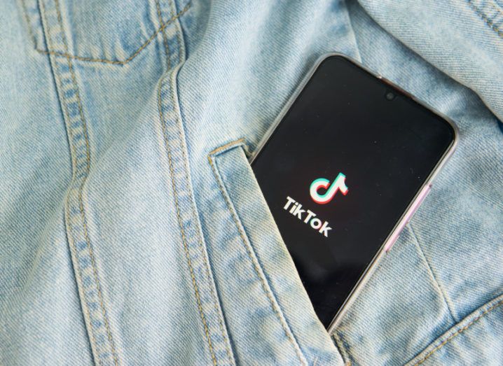 A black smartphone is in the pocket of a denim jacket. The TikTok logo is on the screen of the smartphone.