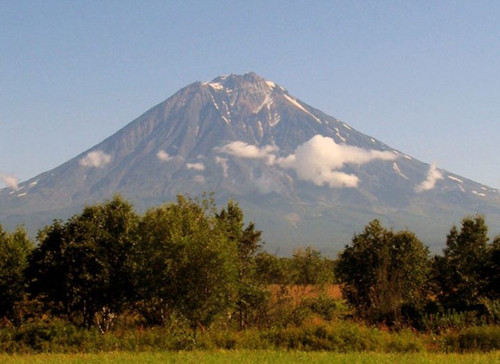 A volcano in the Kamchatka Peninsula is pictured. The volcano is in the distance from the photographer, who is in a field. There are some cirrus clouds visible halfway up the volcano but otherwise the sky is clear. There are some trees between the photographer and the base of the volcano, obscuring the view.