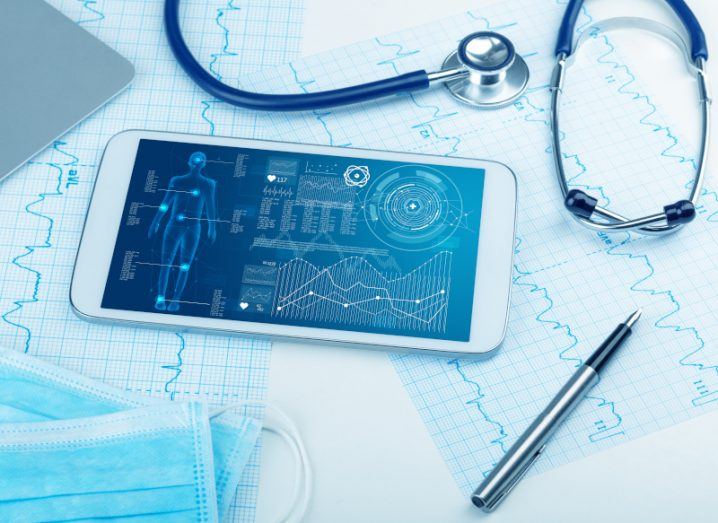 A stock image of a tablet displaying healthcare data beside a stethoscope, surgical masks, and ECG printouts.