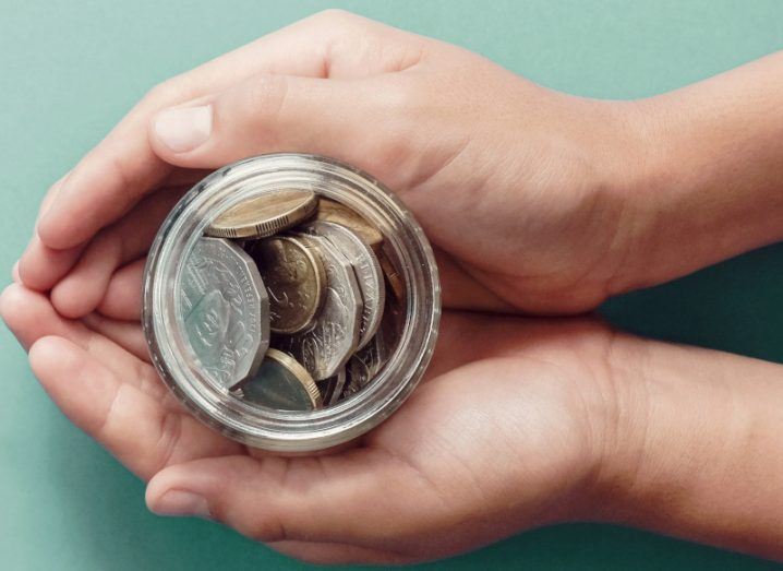 Pair of hands holding a jar of coins against a light blue background.