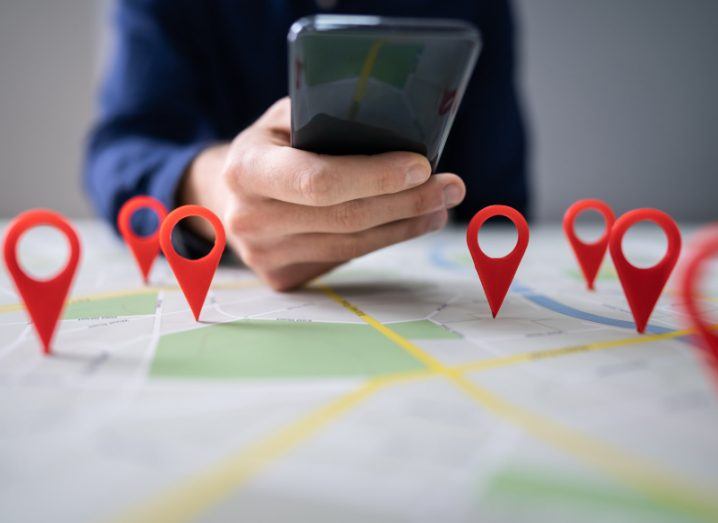 Stalkerware concept: A physical map laid out in front of a person holding a phone. The map has 3D red pins standing up on various locations on the map.