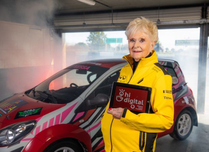 Photo of an older woman in yellow racing gear holding a tablet that reads "Hi Digital". There is a race car in the background.