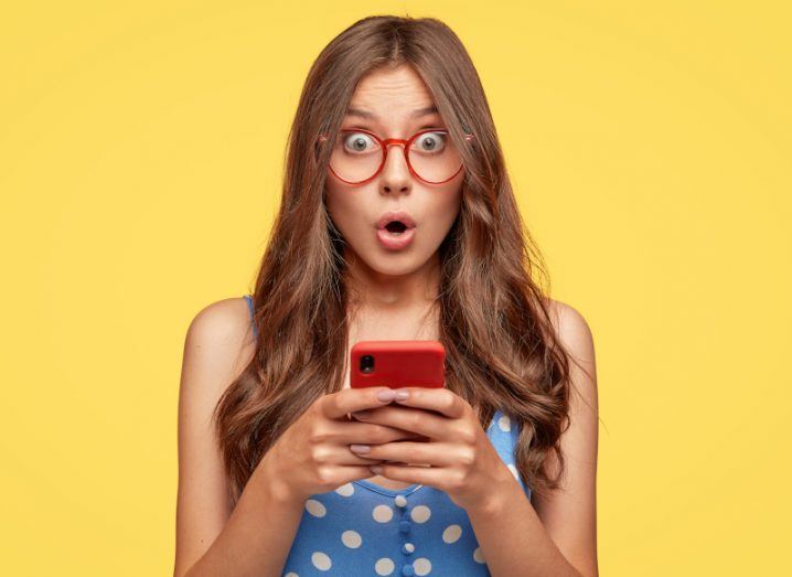 Woman holding a mobile phone in her hands and looking surprised against a yellow background.