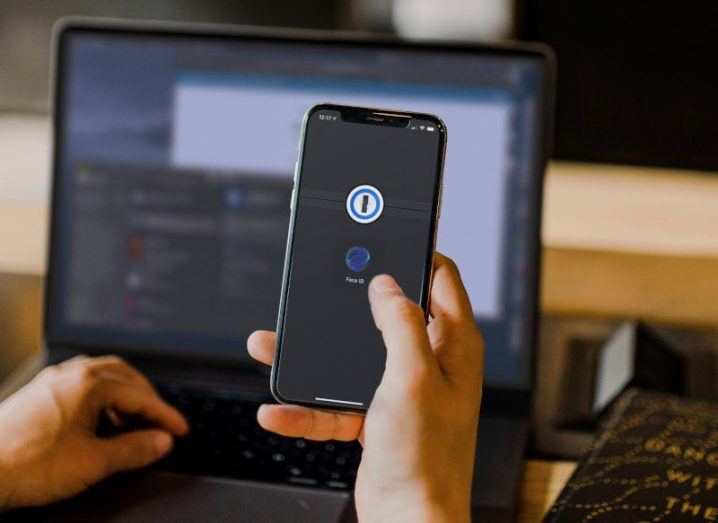 A person holding a phone up with the 1Password logo on the screen. Behind that on the desk is a laptop.