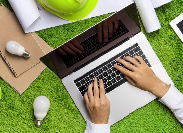 Business person's hands typing on a laptop against a background of fake green grass covered in light bulbs, papers and recycled notebooks.