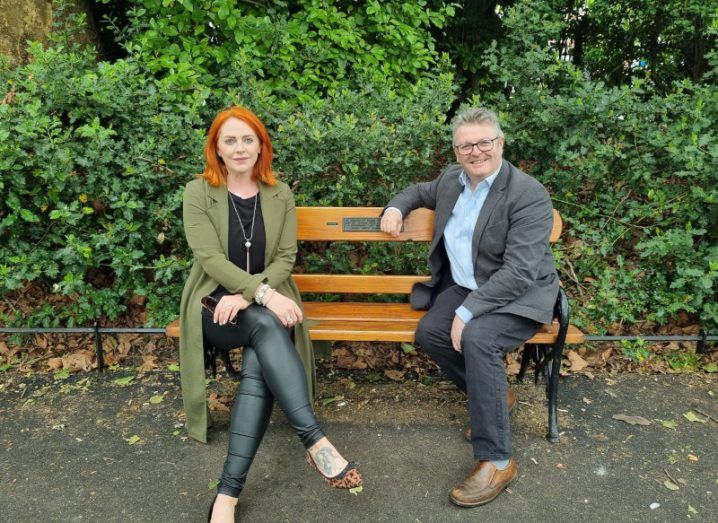 A red-haired woman in black leggings and a long khaki coat sits on a bench alongside a man in a grey suit and pale blue shirt.