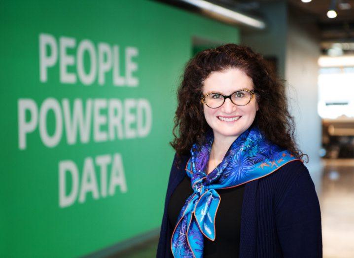 A woman wearing a blue scarf and glasses smiles at the camera. Behind her there is a green wall with white writing that says ‘people powered data’.