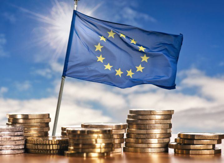 Euro coins on a table with the EU flag behind it, a blue sky with the sun is in the background with some clouds in the distance.