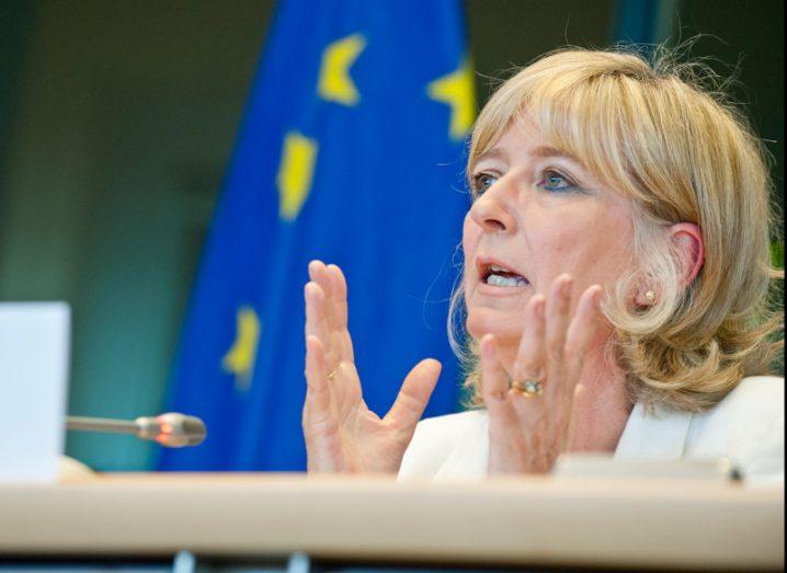 Close-up of Emily O'Reilly talking while seated in the EU Parliament. EU flag in the background.