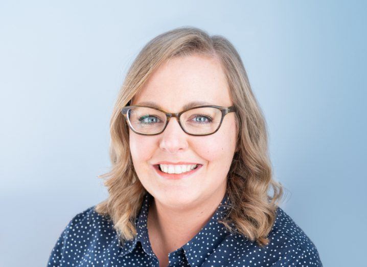 Laurie Winkless, a woman with blonde hair and glasses smiles at the camera against a blue background.