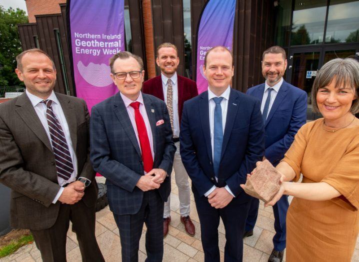 Five men and one woman standing together in front of a building. The woman is holding a brown rock and there are signs with "Northern Ireland Geothermal Energy Week" written on them.
