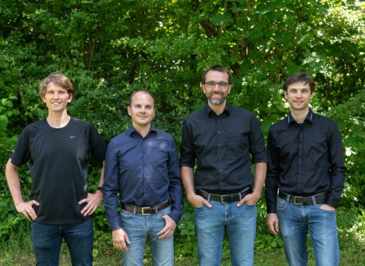 Four men standing side by side smiling at the camera with trees behind them. They are four co-founders of Planqc.