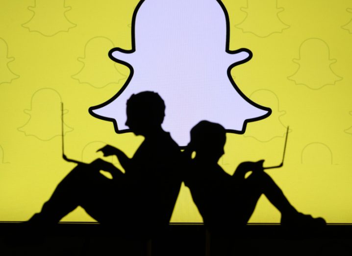 Two silhouettes of people sitting back to back using the web on laptops with Snapchat logo in the background on a yellow background.
