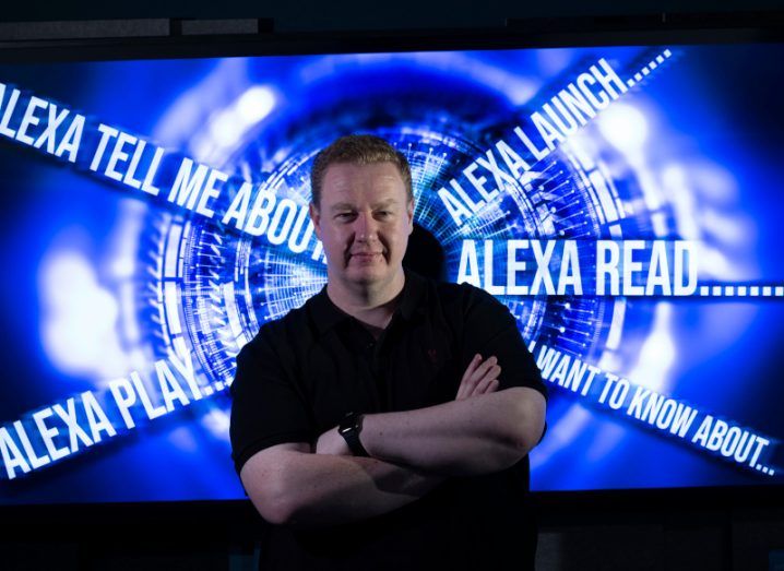 A man in a black T-shirt with her arms folded. Behind him is a graphic of text that shows the beginning of Alexa commands.