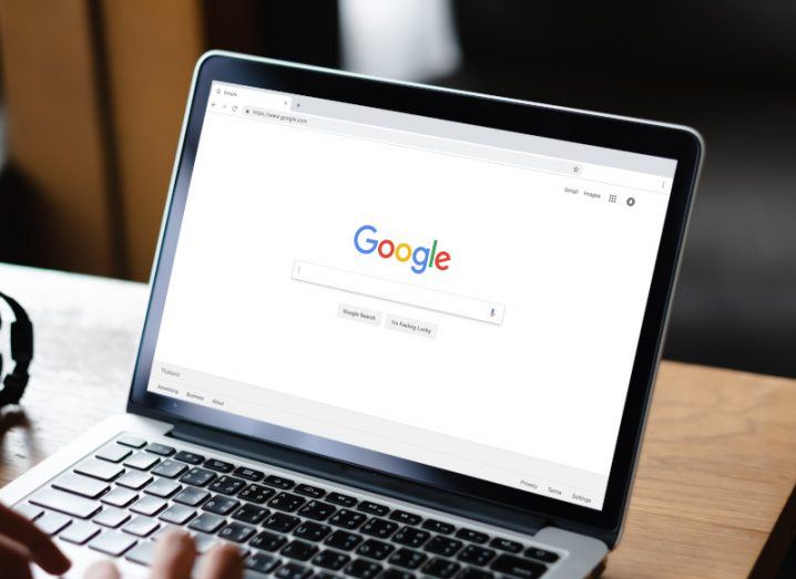 A laptop with the Google Search page on the screen, with a person's hand typing on the keyboard. The laptop is resting on a wooden table.