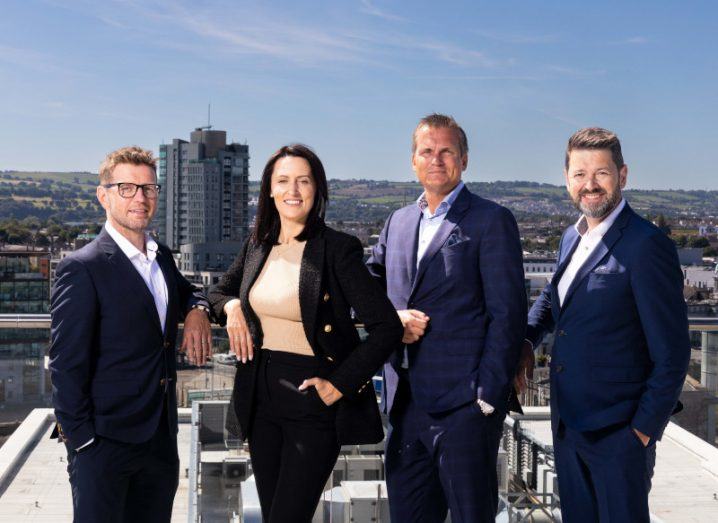 Three men and one woman standing side by side on a roof, with buildings, hills and a blue sky in the background.