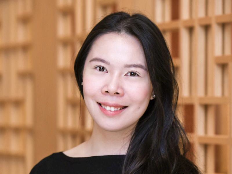 A headshot of Rhea See, smiling in front of a wooden feature wall.