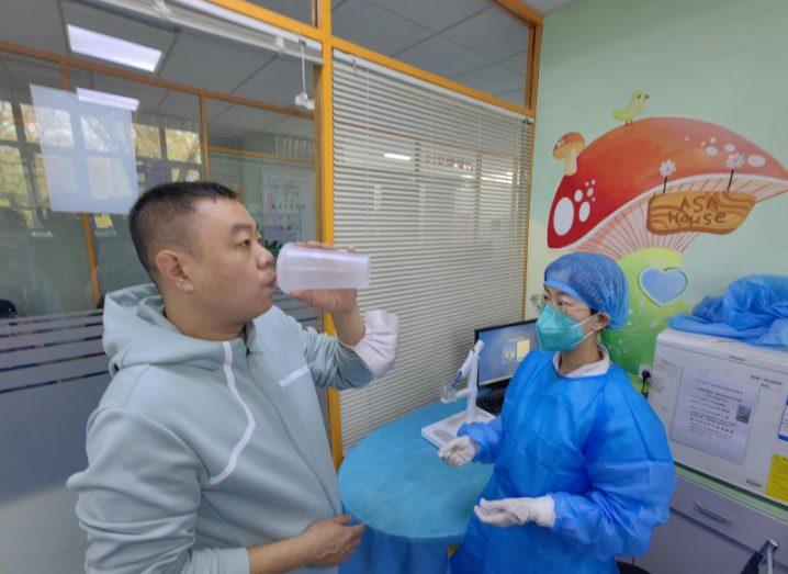 A person holding a cup against his mouth, with a medical worker standing next to him wearing a mask. Used to show the inhaled Covid-19 vaccine being developed by CanSanBio, in partnership with Aerogen.