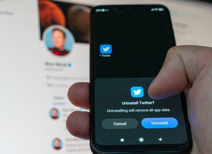 A person is holding up a phone, about to press a button to uninstall Twitter. The Twitter profile of Elon Musk looms in the background.