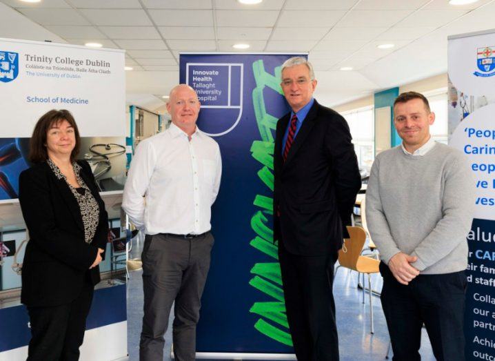 Three men and a woman standing together in the interior of a hospital. There are signs behind them that reference Tallaght University Hospital and Trinity College Dublin.