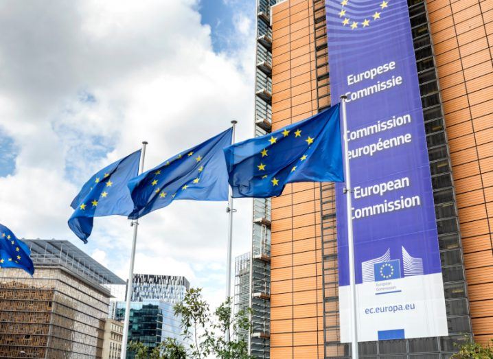 Three EU flags in front of the European Commission headquarters, with a cloudy sky in the background.