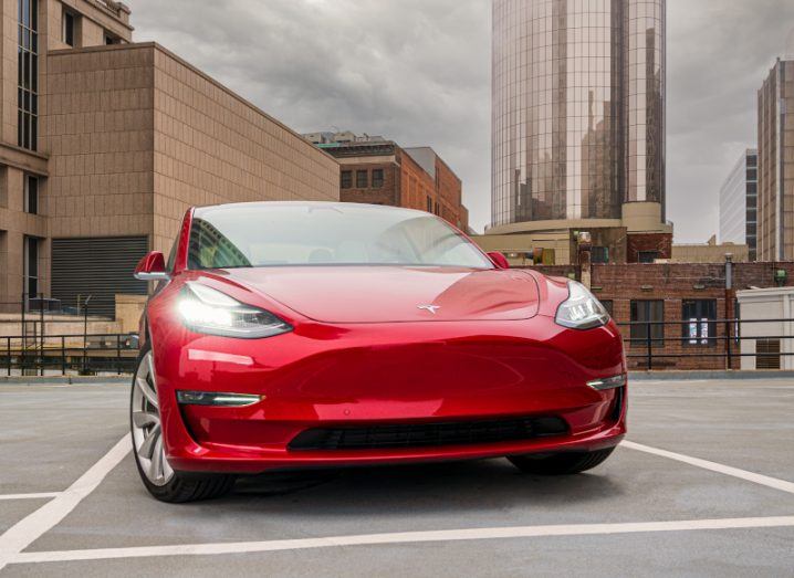 A red Tesla vehicle on a rooftop car park, with buildings and a grey cloudy sky in the background.