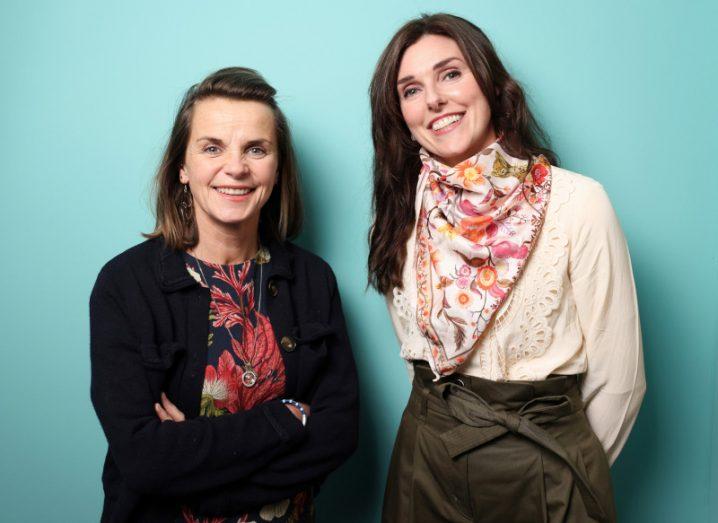 Two women stand in front of an aquamarine background and smile at the camera.