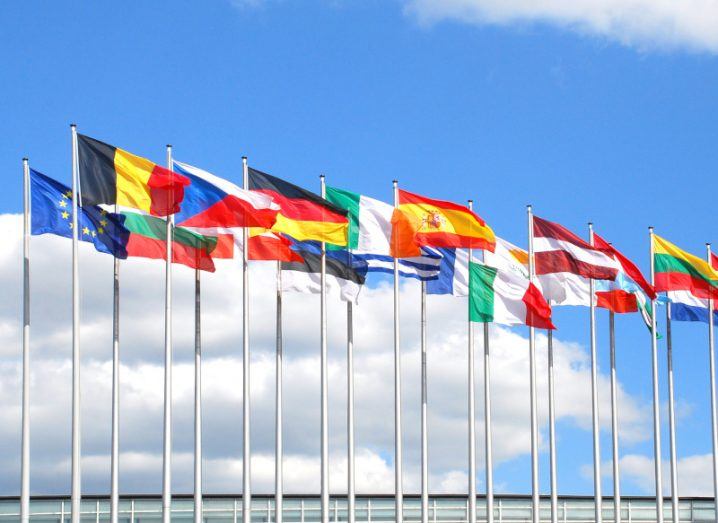 A group of flags including the Irish and EU flag, on flag poles with a blue sky and clouds in the background.